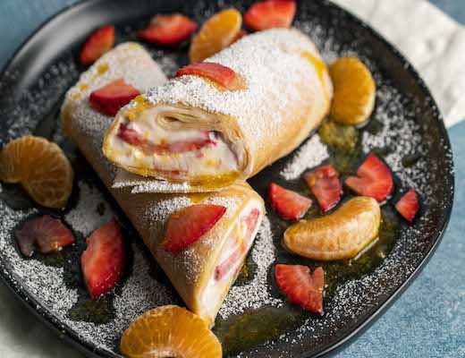 Delicious rolled crepes with strawberries and citrus on a black plate.