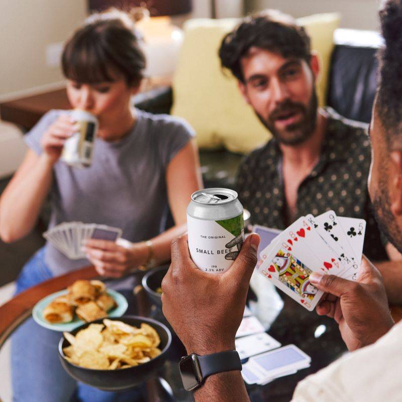 Three young adults drink cans of mid-strength beer while playing cards and eating party food