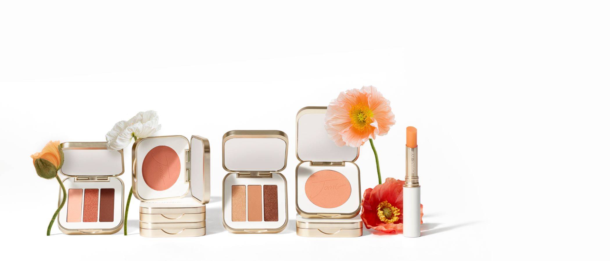 Several jane iredale products including eyeshadow, blush and a lip stain in shades of peach, coral, and copper are lined up with a few flowers against a white background.