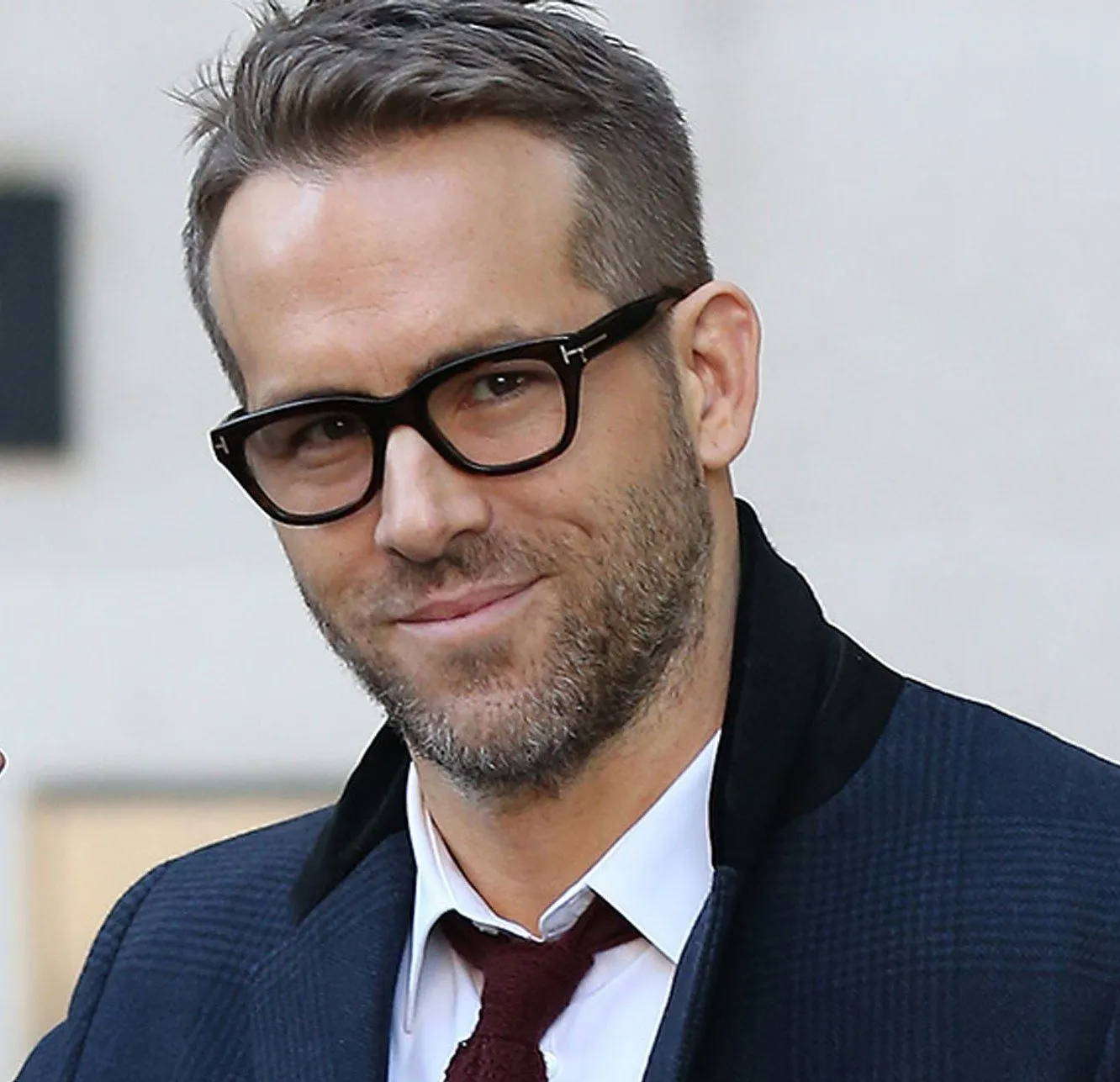 Ryan Reynolds wearing a blue coat, white shirt, red tie and thick frame glasses