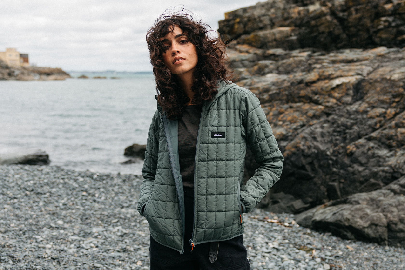 Person in a quilted green jacket standing by the rocky shore.