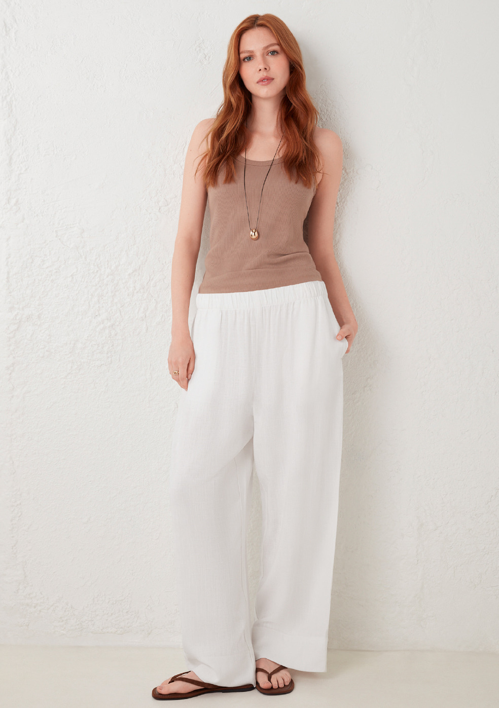 Woman in casual outfit with tank top and wide pants standing against a white wall.