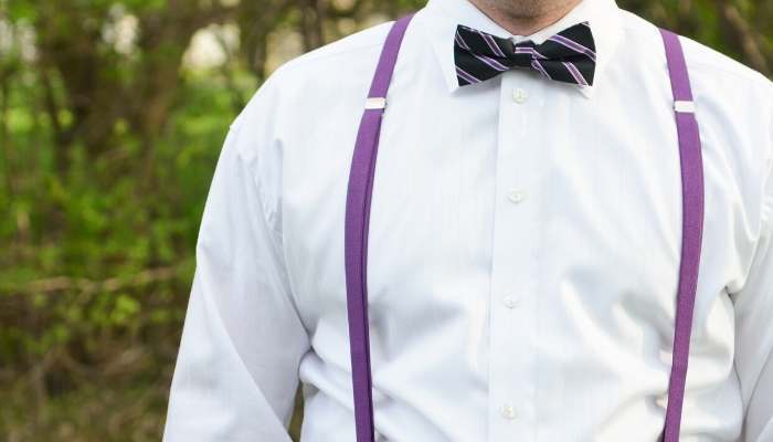 Man wearing purple skinny suspenders, a white shirt and black and purple striped bow tie outside