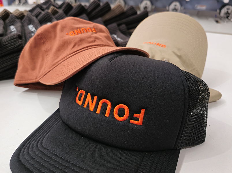 Stack of orange, black and tan 'Found Brewing' trucker caps with embroidered logo on the front, labelled Embroidery