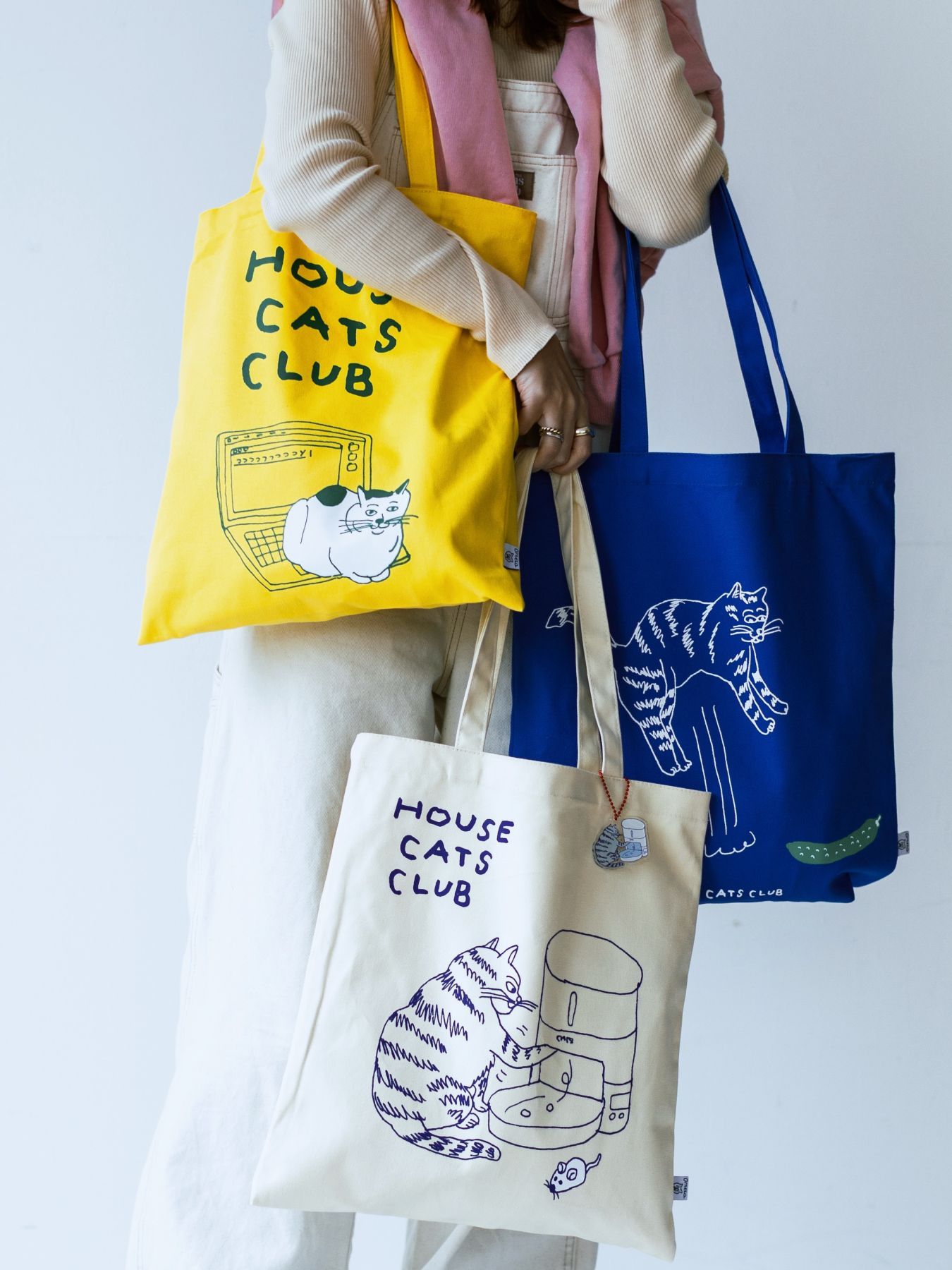 Person holding three tote bags featuring cat-themed designs.
