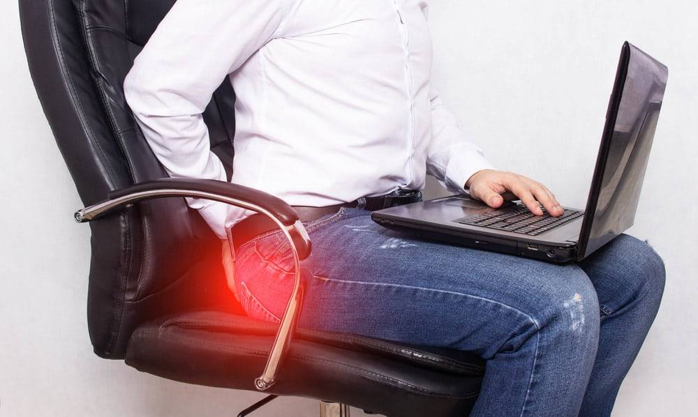 This is a picture of a man sitting on a chair with his laptop on his lap holding his lower back in pain.