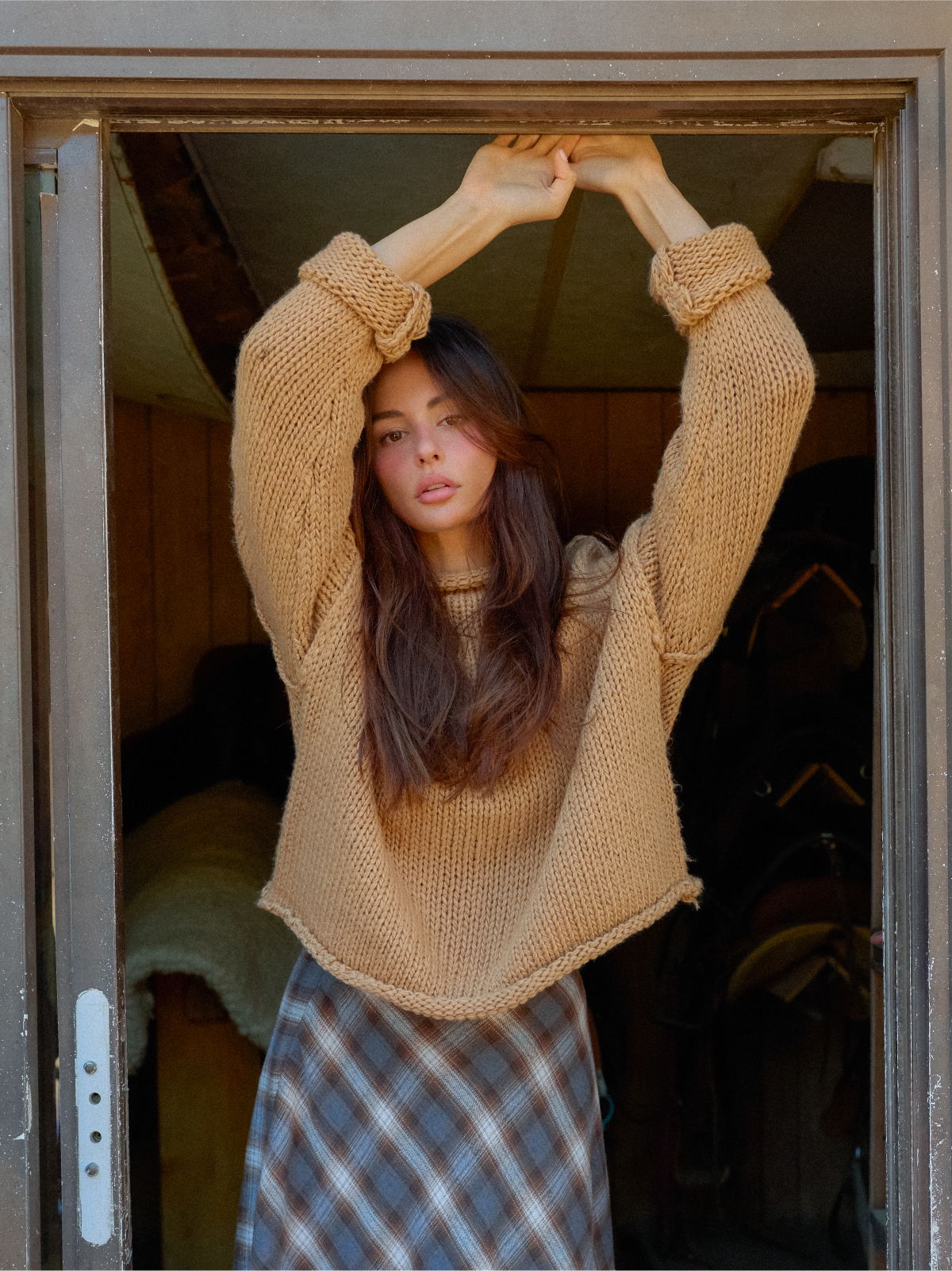 Young woman stretching in doorway, wearing a cozy sweater and plaid skirt.