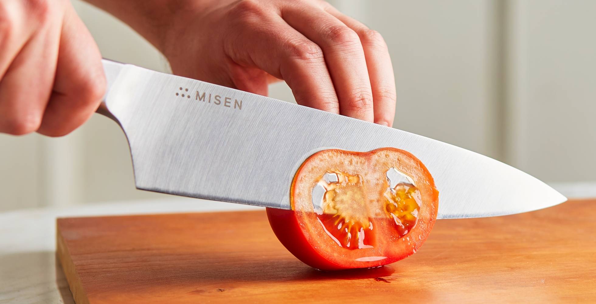 Hands use a blue Misen Chef’s Knife to cut a nearly transparent slice of tomato.