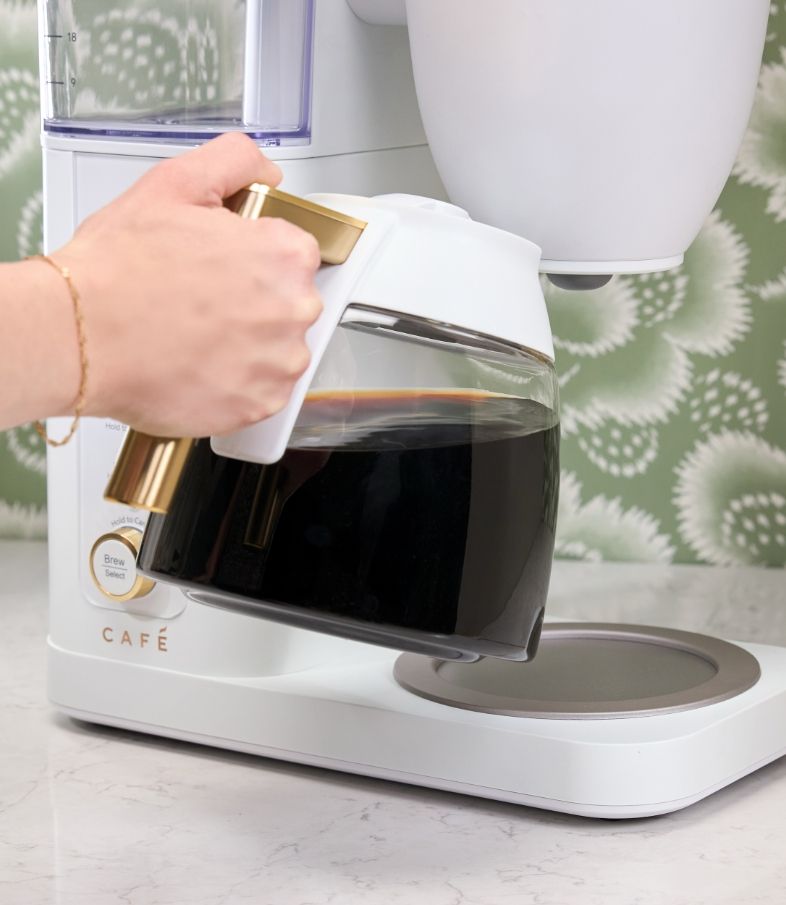 Hand holding a coffee pot over a coffee maker on a countertop.