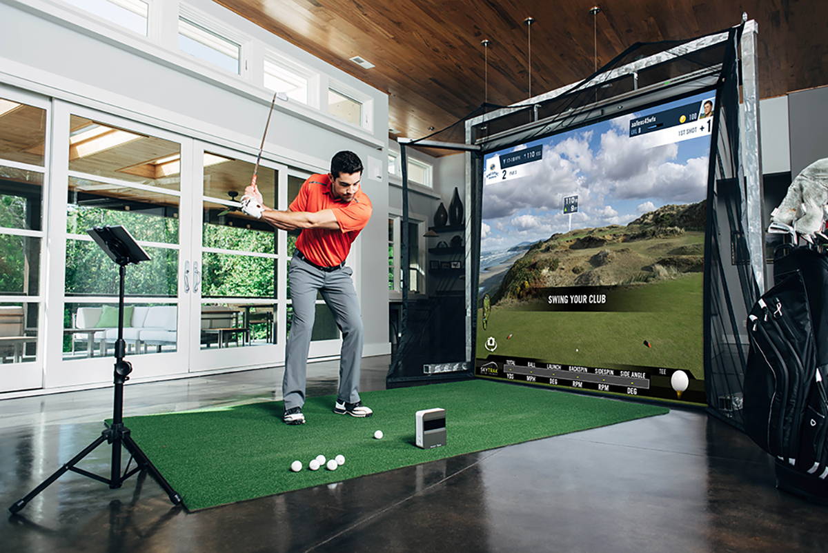 A man starting a swing in a home golf simulator with SkyTrak golf launch monitor, impact screen, golf mat, and tablet on a stand