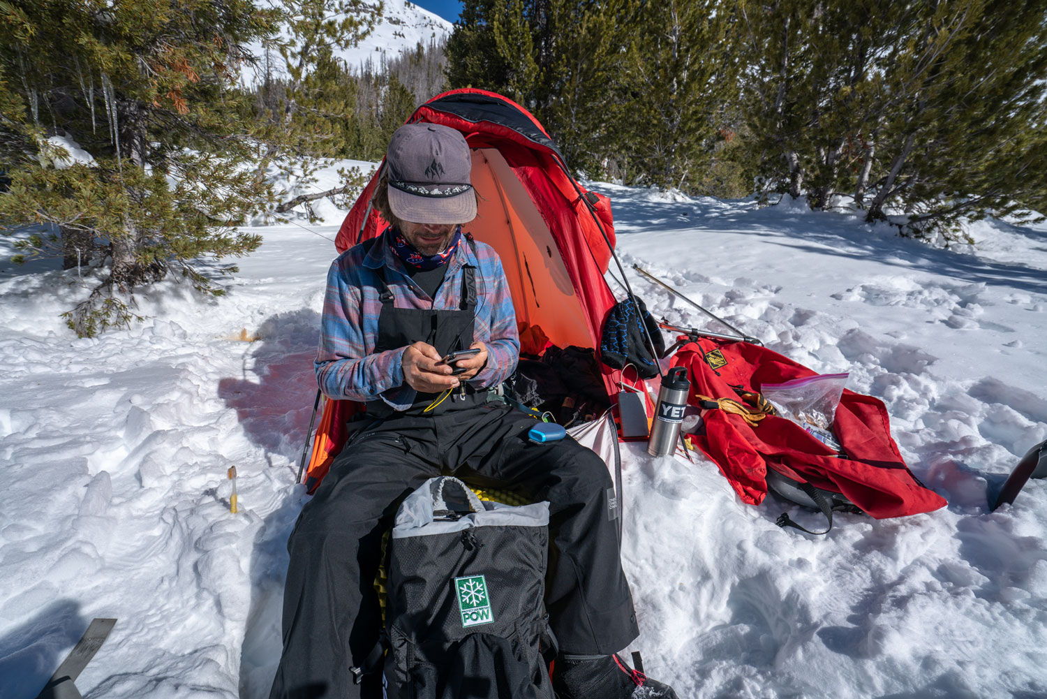Person sitting near a tent in the snow, using a device.