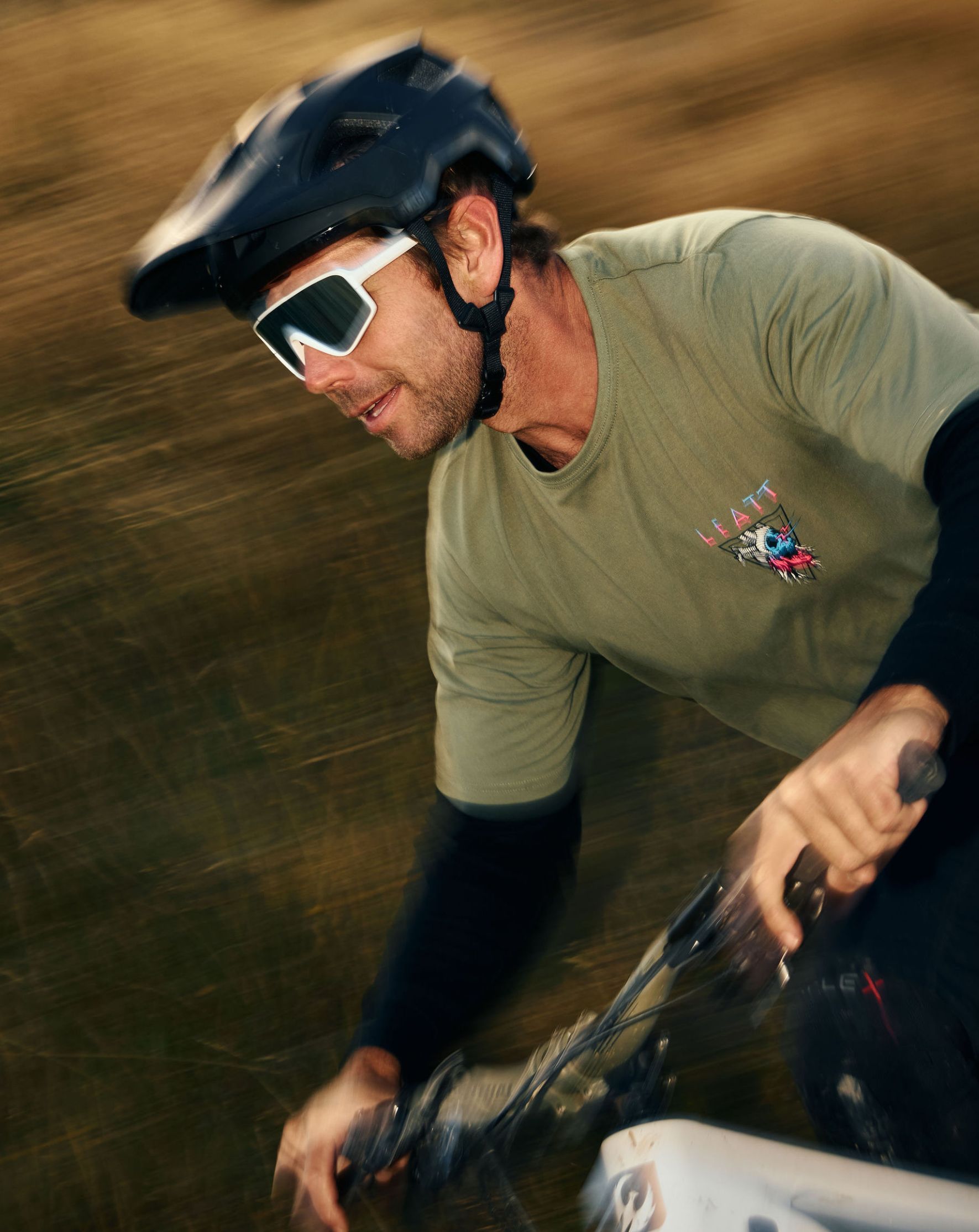A cyclist wearing a helmet and sunglasses rides through a blurred background.