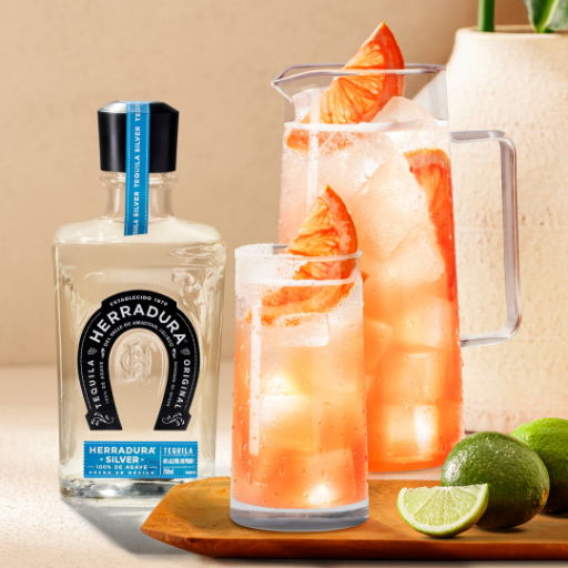 Pitcher and glass of paloma cocktail with limes and a bottle of Herradura silver tequila.