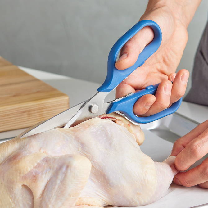 A chef using Blue Misen Kitchen Shears to remove the spine from a raw chicken.