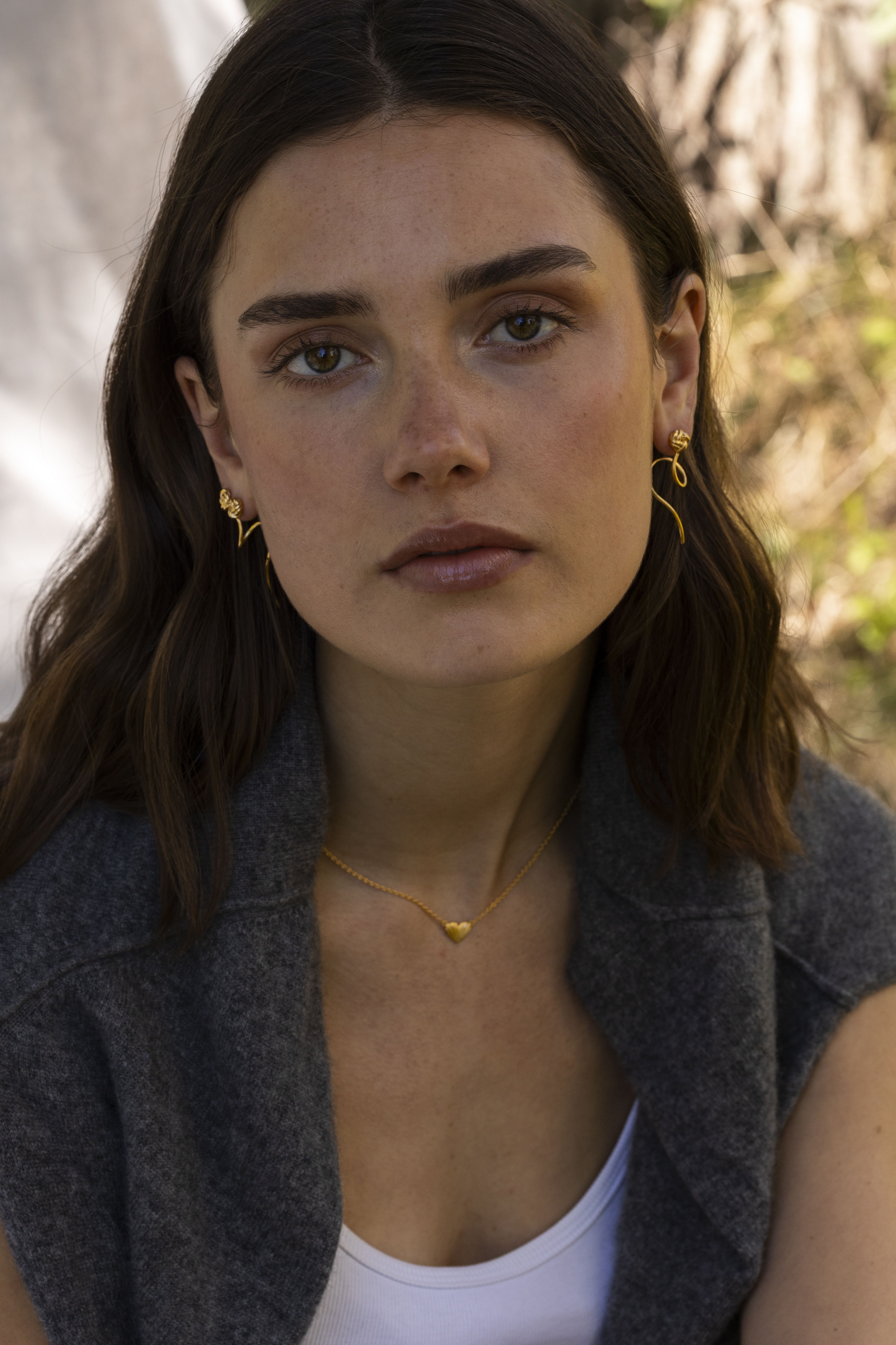 Young woman with brown hair wearing earrings and a necklace, looking directly at camera.
