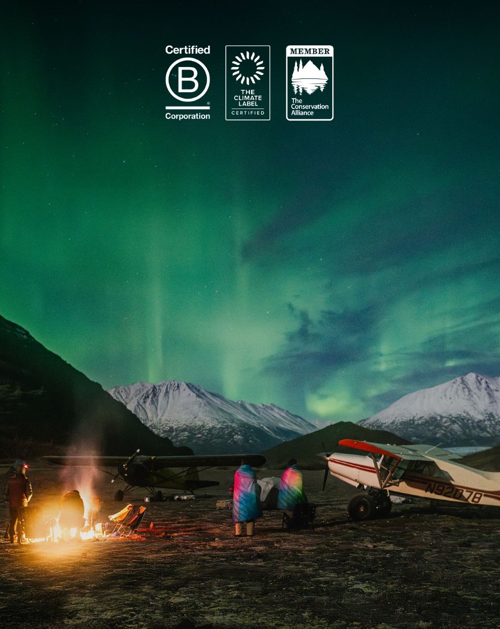 People gathered around a fire under the northern lights with mountains in background.