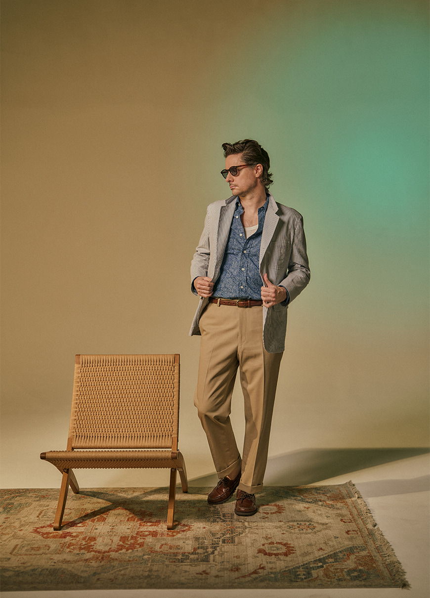 Man in stylish outfit stands next to a chair on a patterned rug.