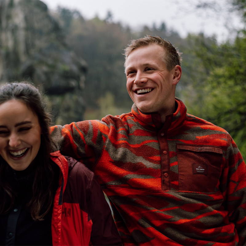 Man & Woman both wearing red Helly Hansen fleece jackets in woods.