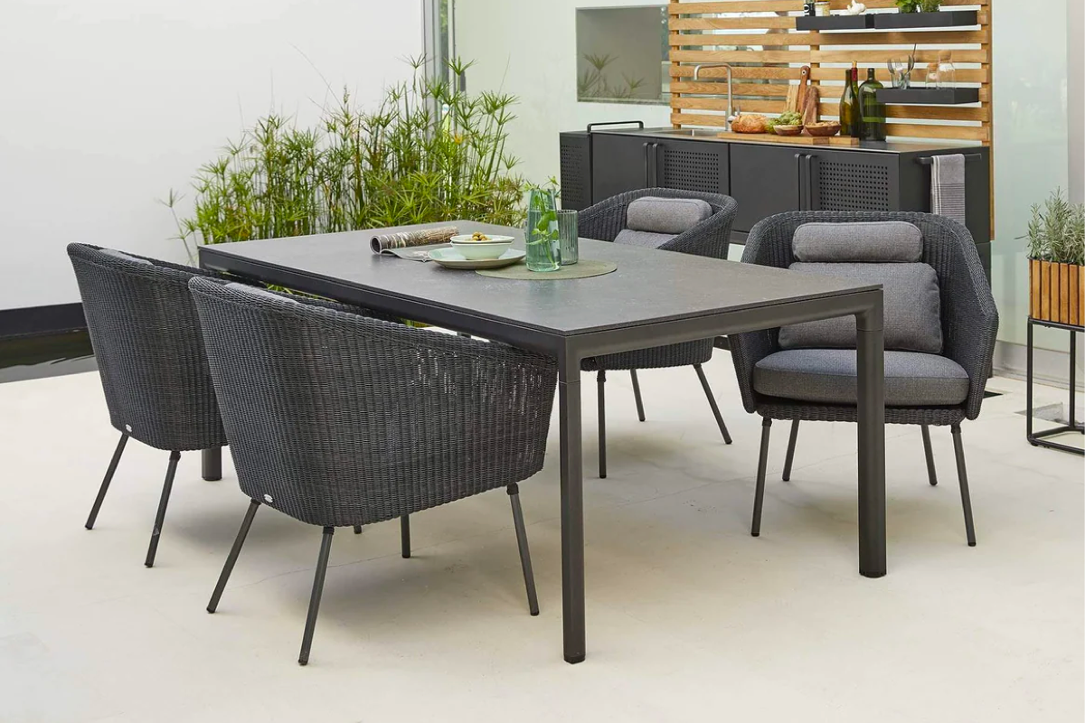 A dark grey outdoor dining table with plush woven grey seats on a patio with grasses and outdoor kitchen in the background. 