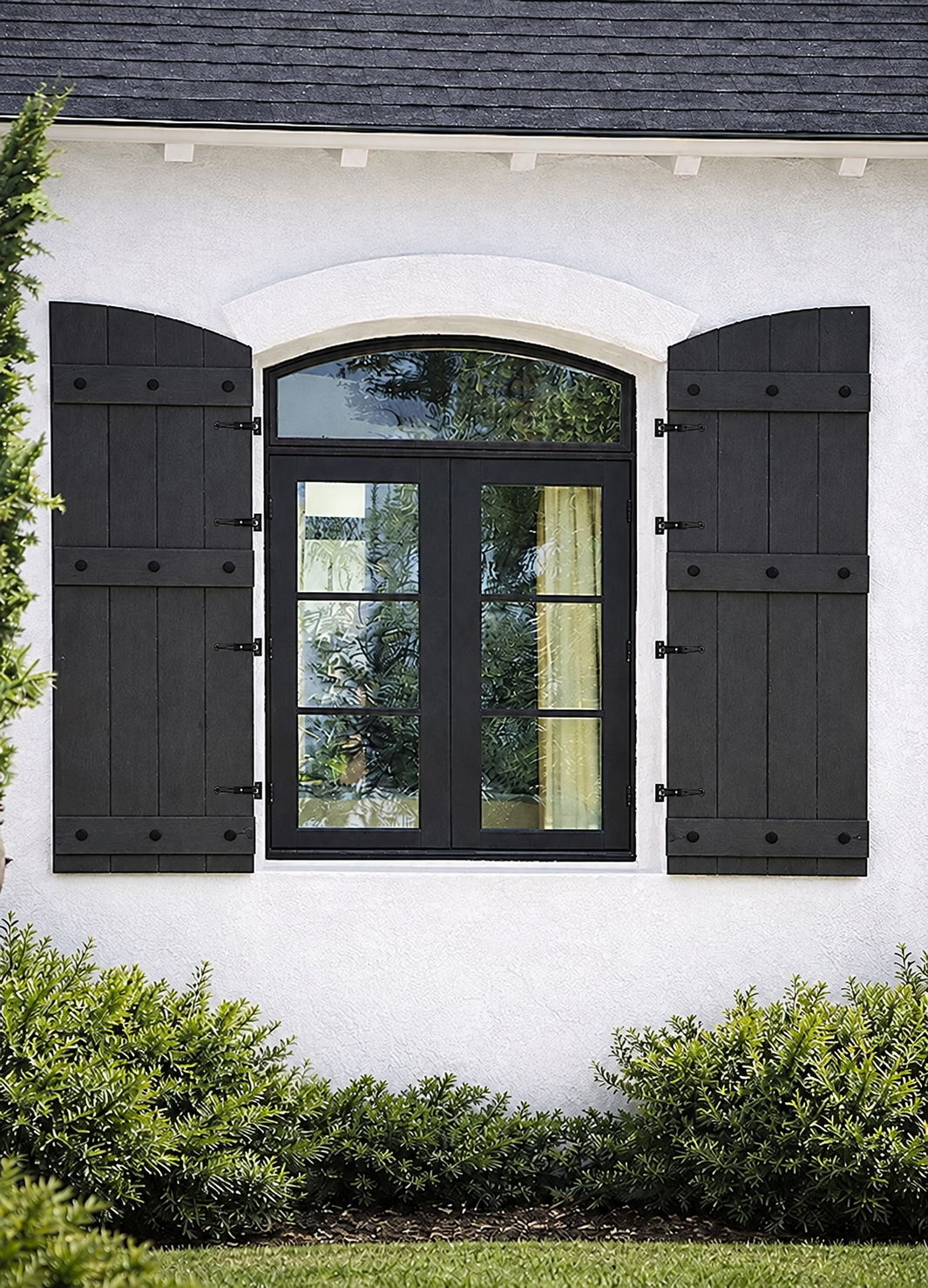 Black window with arched shutters on a white wall surrounded by greenery.