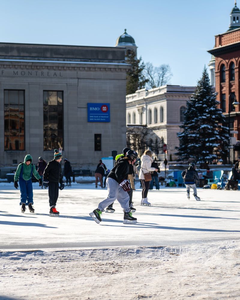People ice skating in a snowy outdoor rink surrounded by buildings.