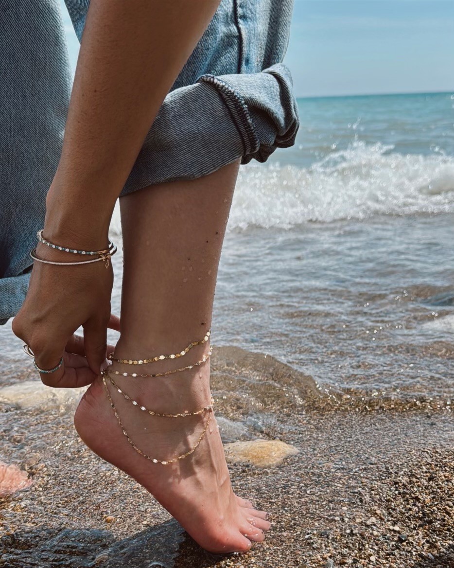 A person adjusting anklets while standing on a beach near the water.