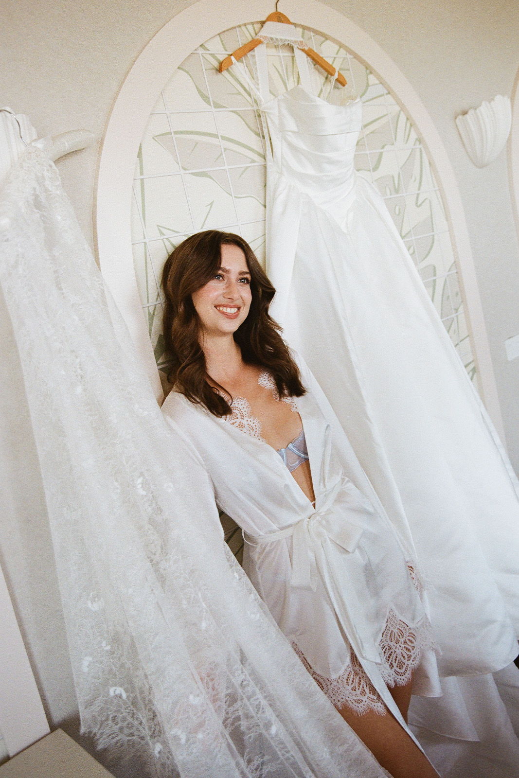 Smiling woman in a white robe beside wedding dresses and veil.