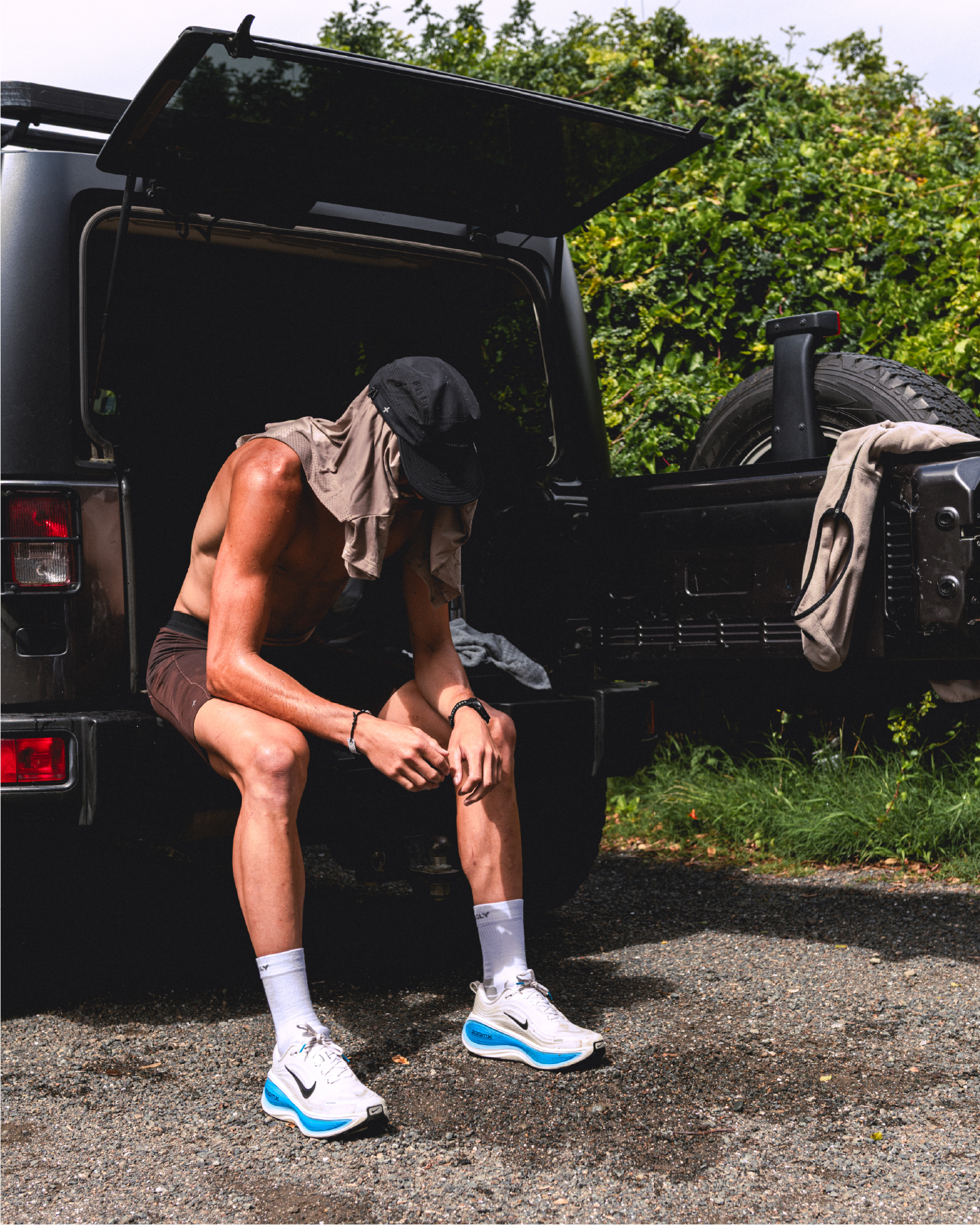 A shirtless man sitting by a vehicle, looking contemplative outdoors.