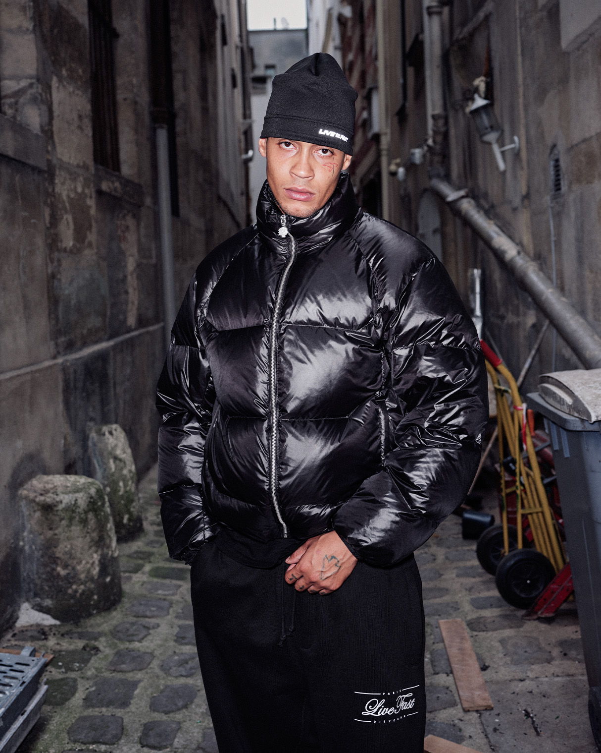 Person in black puffer jacket and hat standing in an alleyway.
