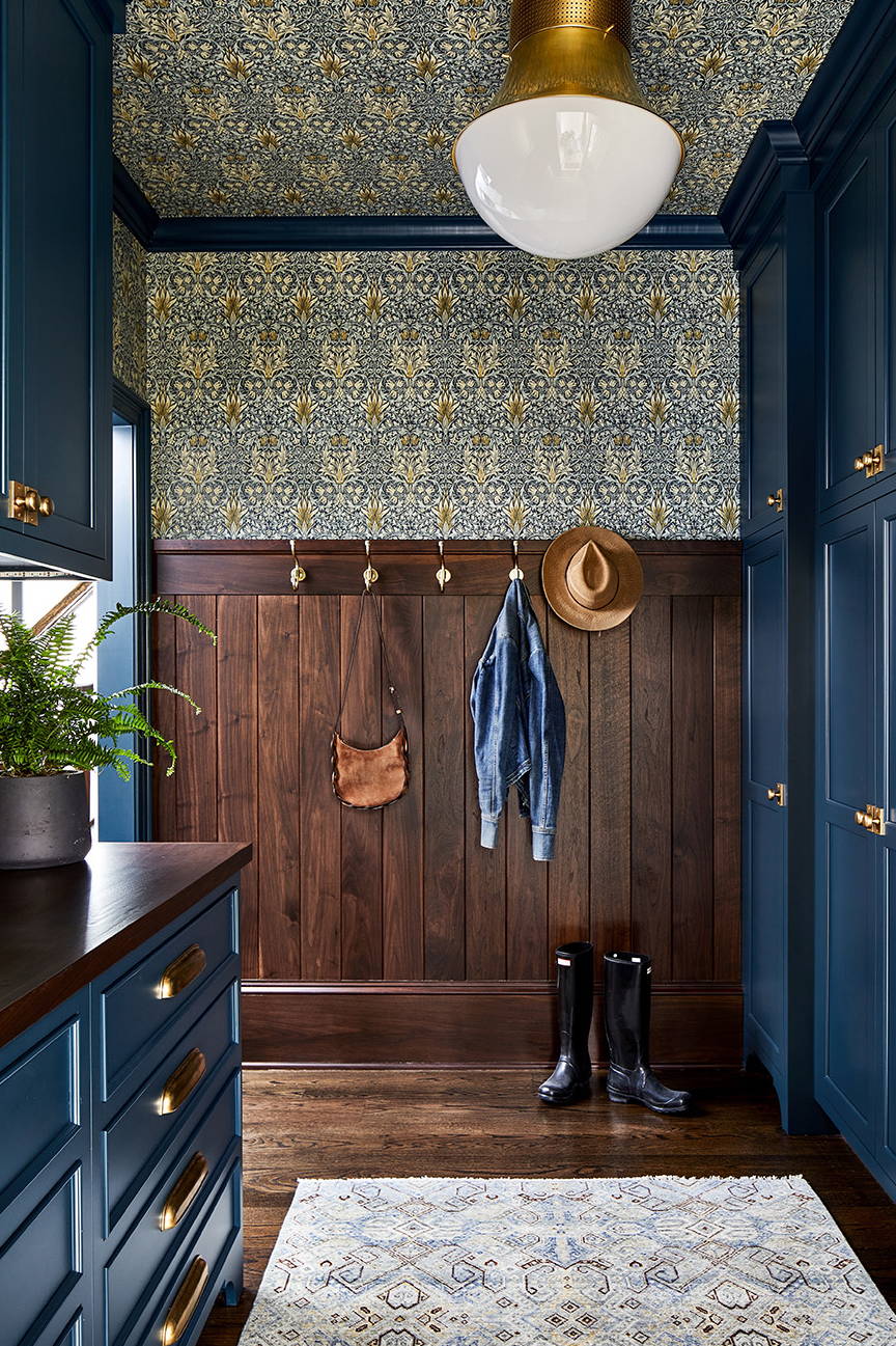 Traditional mudroom design by Elizabeth Reich Interiors with navy cabinetry, brass hardware, wood panelling, floral wallpaper, and wooden flooring