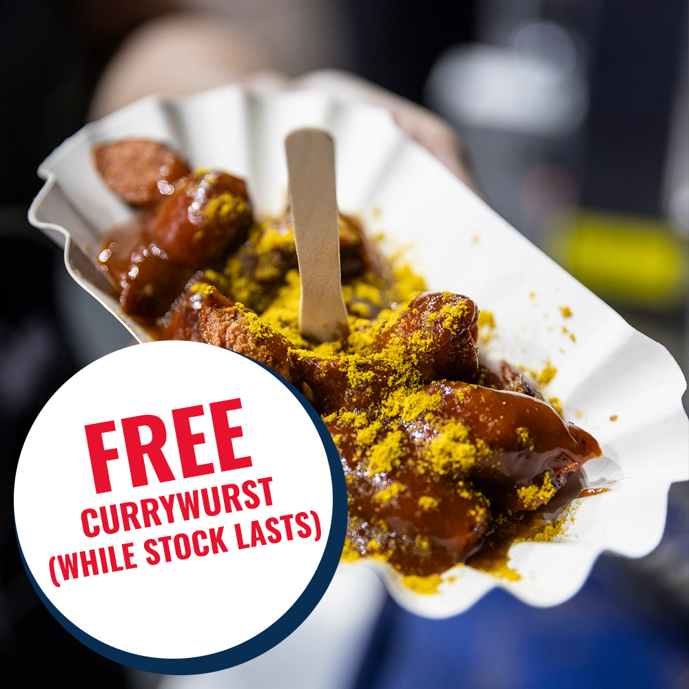 A hand holding currywurst with a promotional sign.