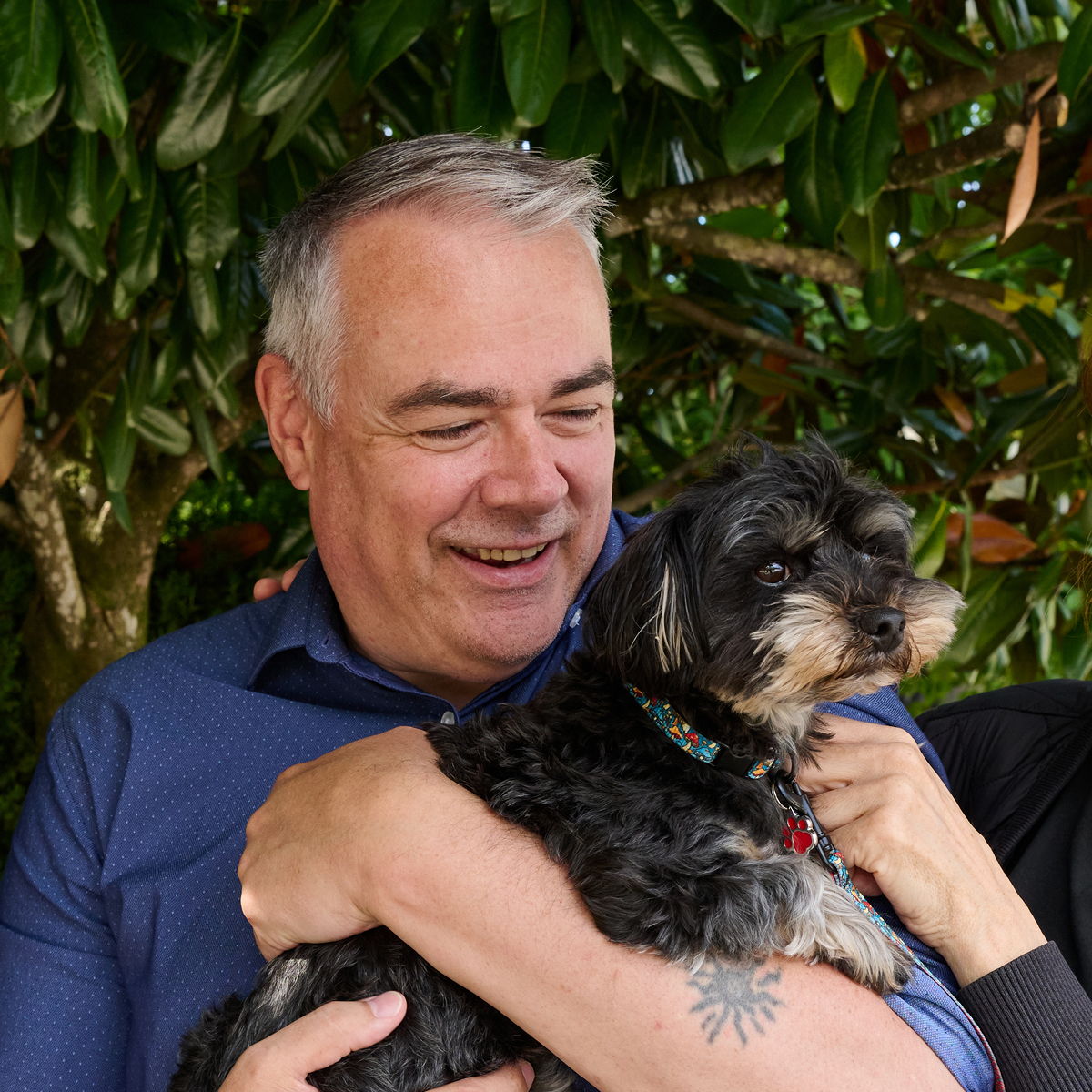 Smiling man holding a small dog with greenery in the background.