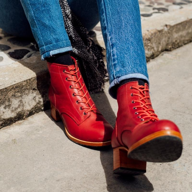Close-up lifestyle view of a person wearing the handcrafted Adelante Elena leather lace-up ankle boots in vibrant red, paired with blue jeans and sitting outside on a sunny day.