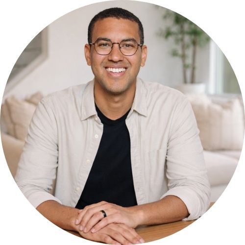 Smiling person with glasses sitting indoors, wearing a light shirt and black shirt.