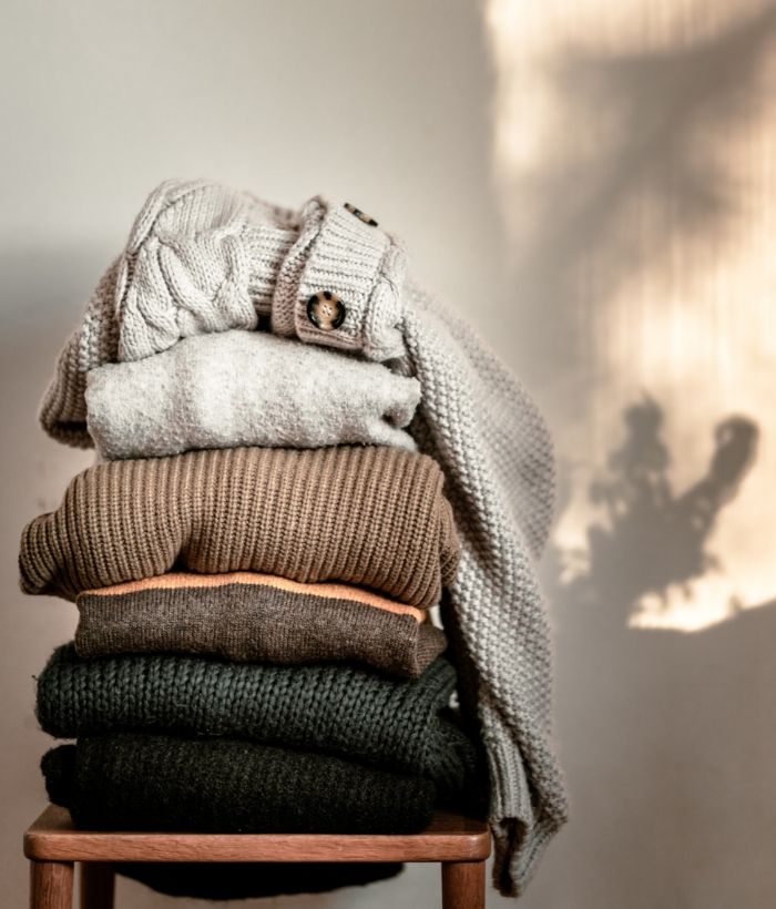 A stack of folded sweaters in neutral colors on a wooden stool.