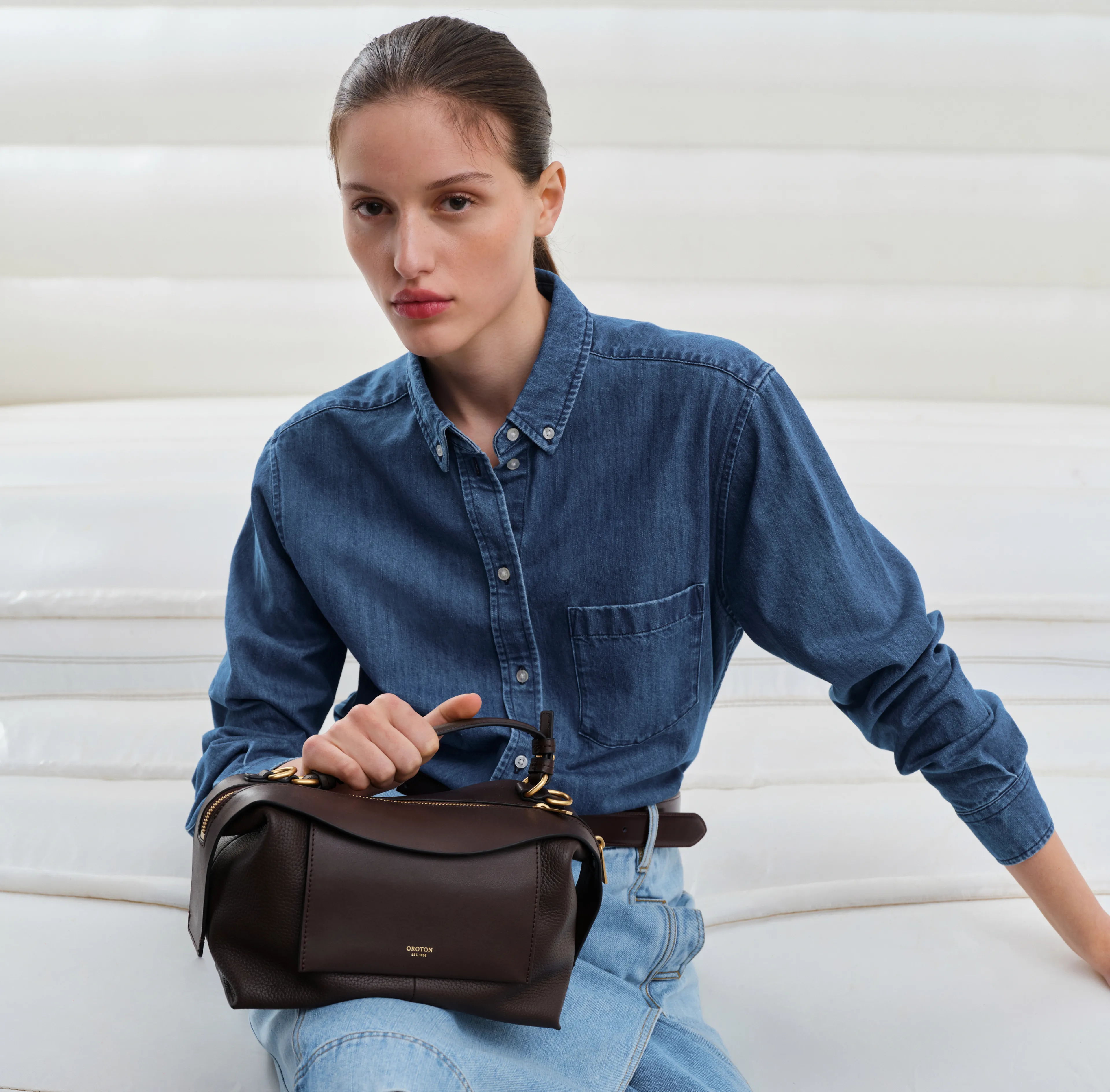Woman in a denim shirt holding a brown handbag, sitting against a white background.