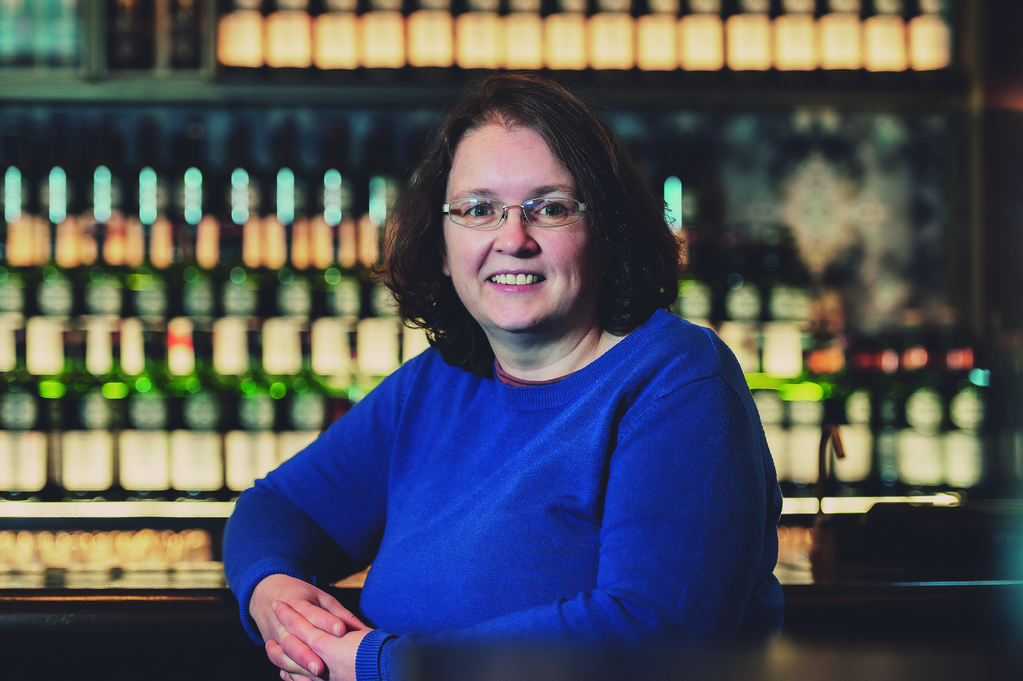 Smiling woman in a blue sweater poses in front of a bar shelf.