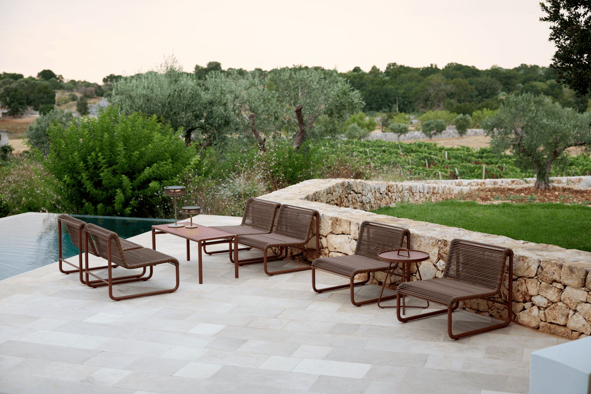 Durable outdoor lounge chairs and side tables arranged beside a pool on a natural stone patio with olive trees