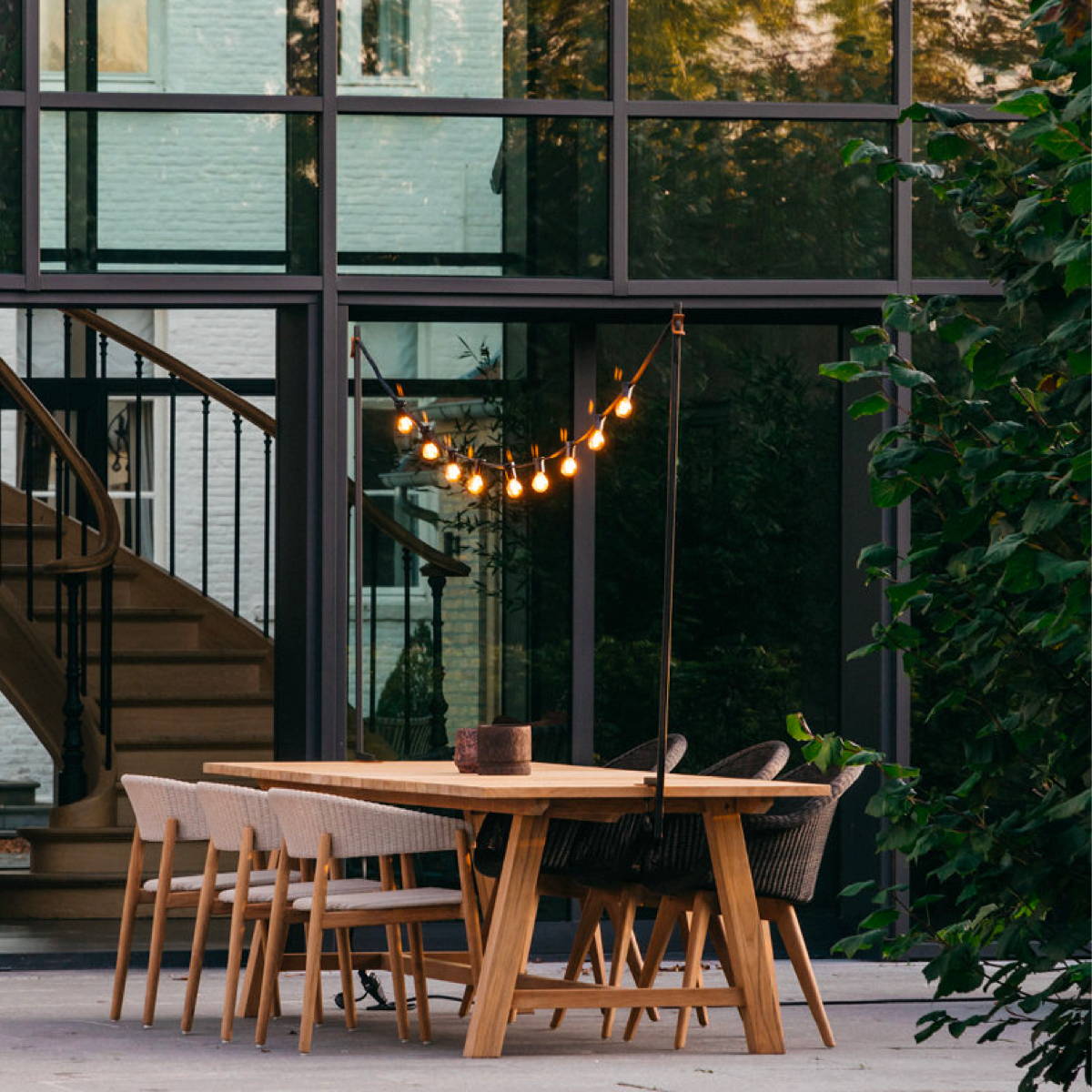 Six Mona dining chairs set at a wooden outdoor dining table under a string of lights.
