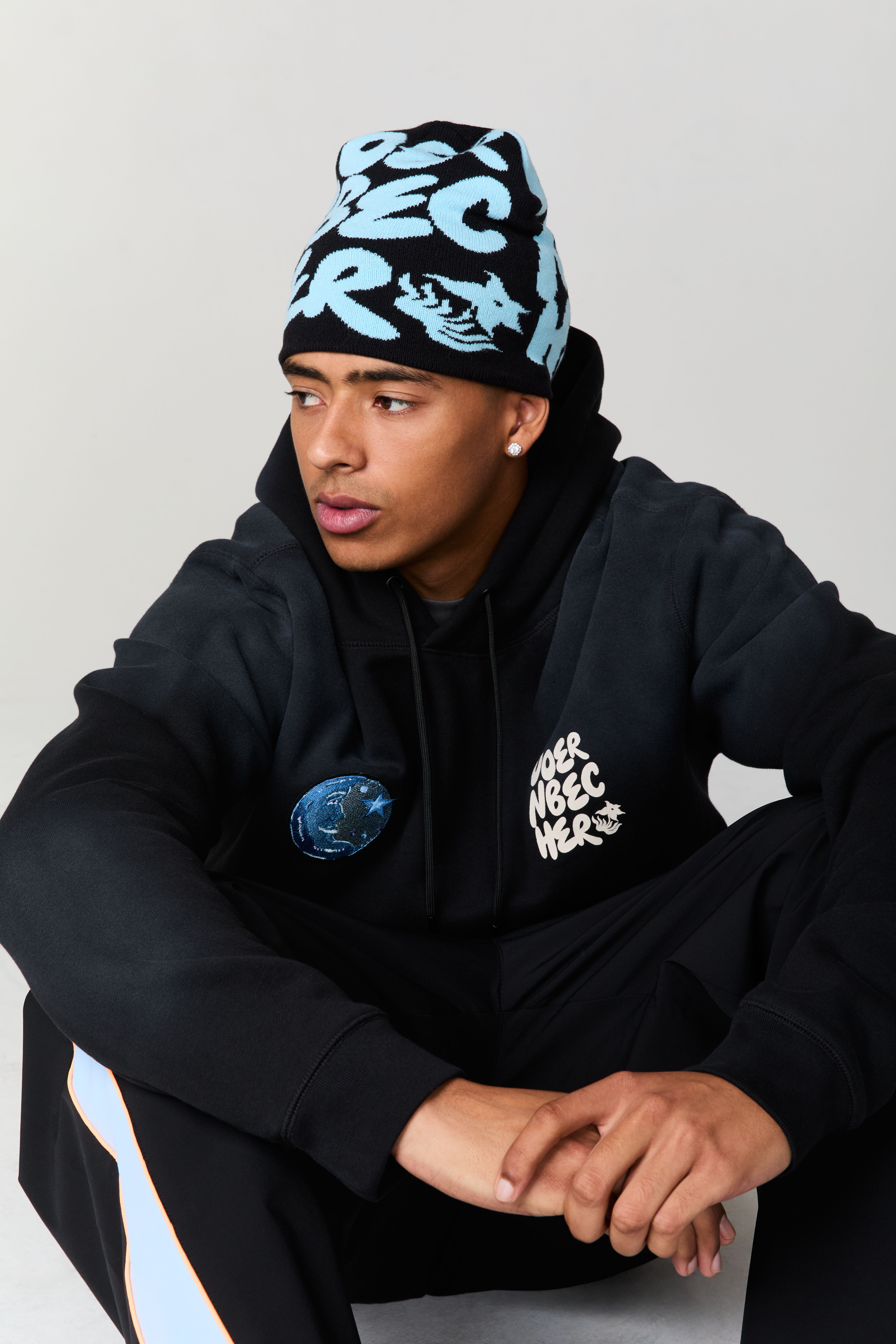 Young man in black hoodie and beanie, sitting against a neutral background.