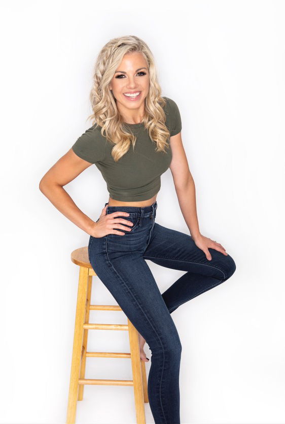 A smiling person with blonde hair sitting on a stool, wearing a green top and jeans.