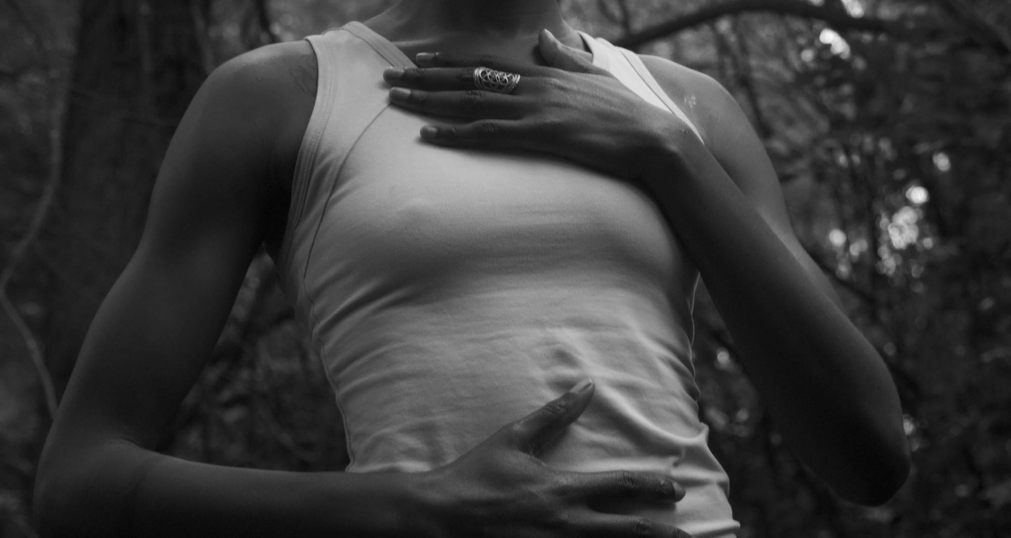 A woman wearing a white shirt touches her chest and abdomen as she stands in a forest.