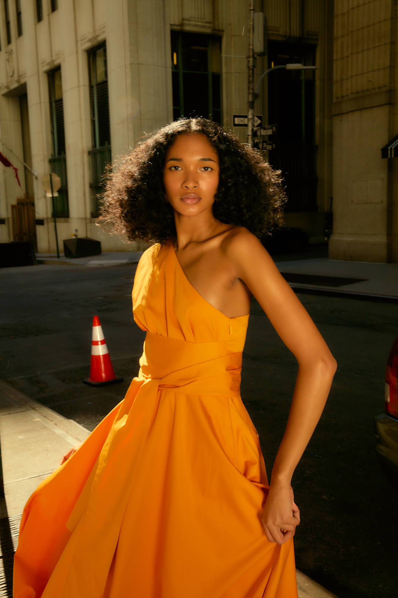 A woman in an orange dress poses outdoors with city buildings in the background.