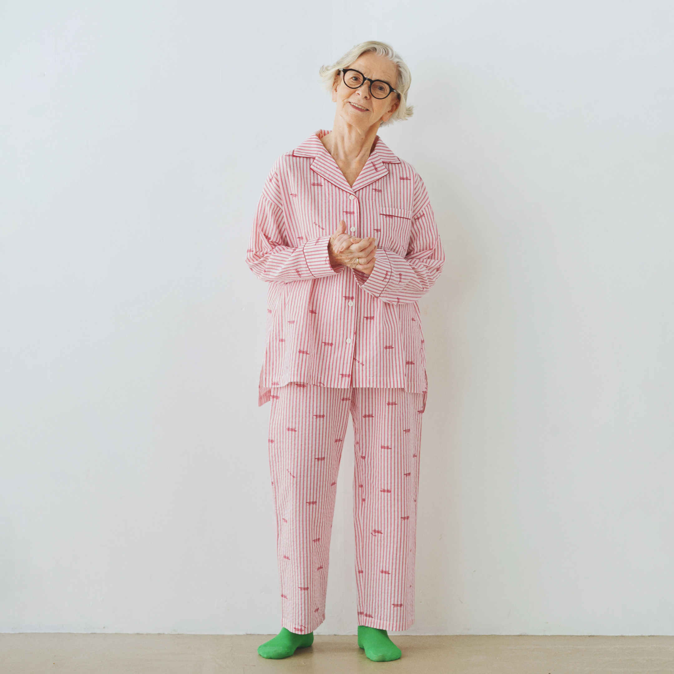 An elderly woman in pink pajamas and green slippers standing against a white wall.