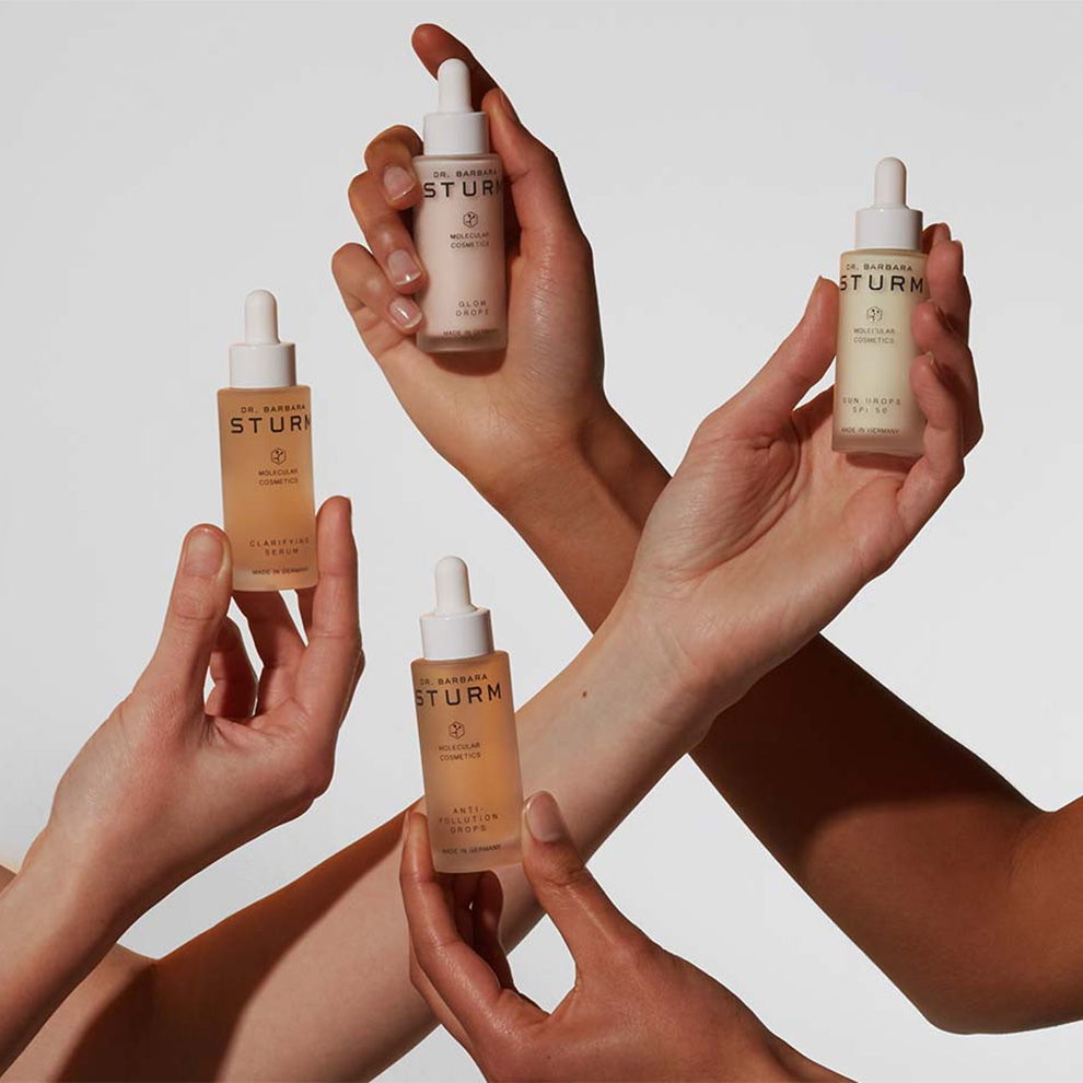 Four hands holding different bottles of skincare serums against a neutral background.