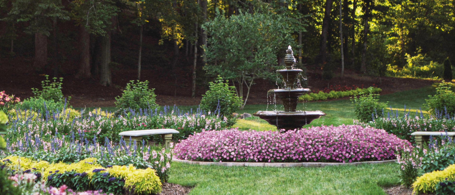 Water fountain surrounded my pink annuals planted in a garden with a bench surrounded by more annuals