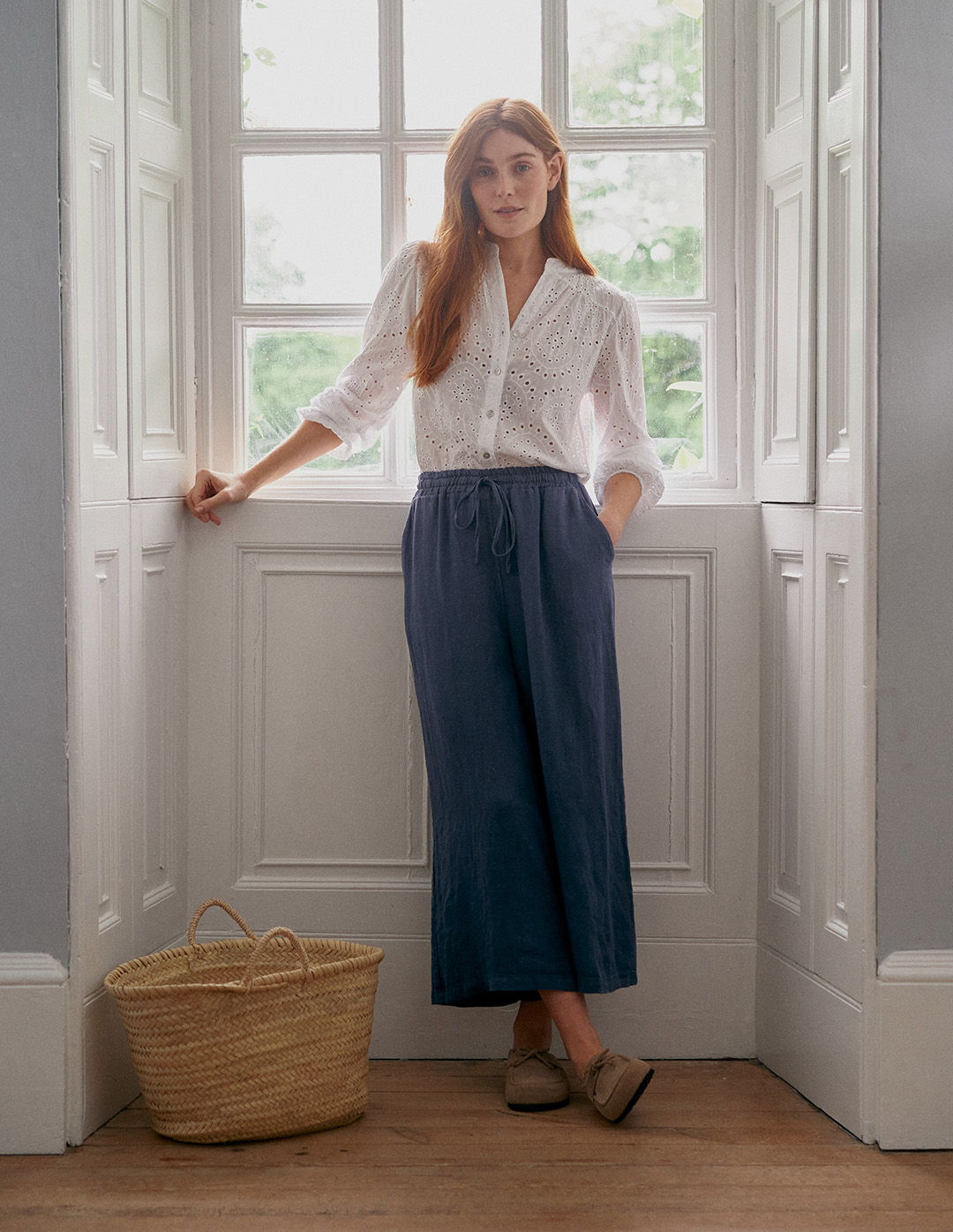 Woman standing in a window with a white broderie anglaise top and blue cropped trousers