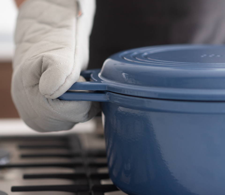 Hand with gray oven mitt firmly gripping the handles of a Blue Misen Dutch Oven with Grill Lid.