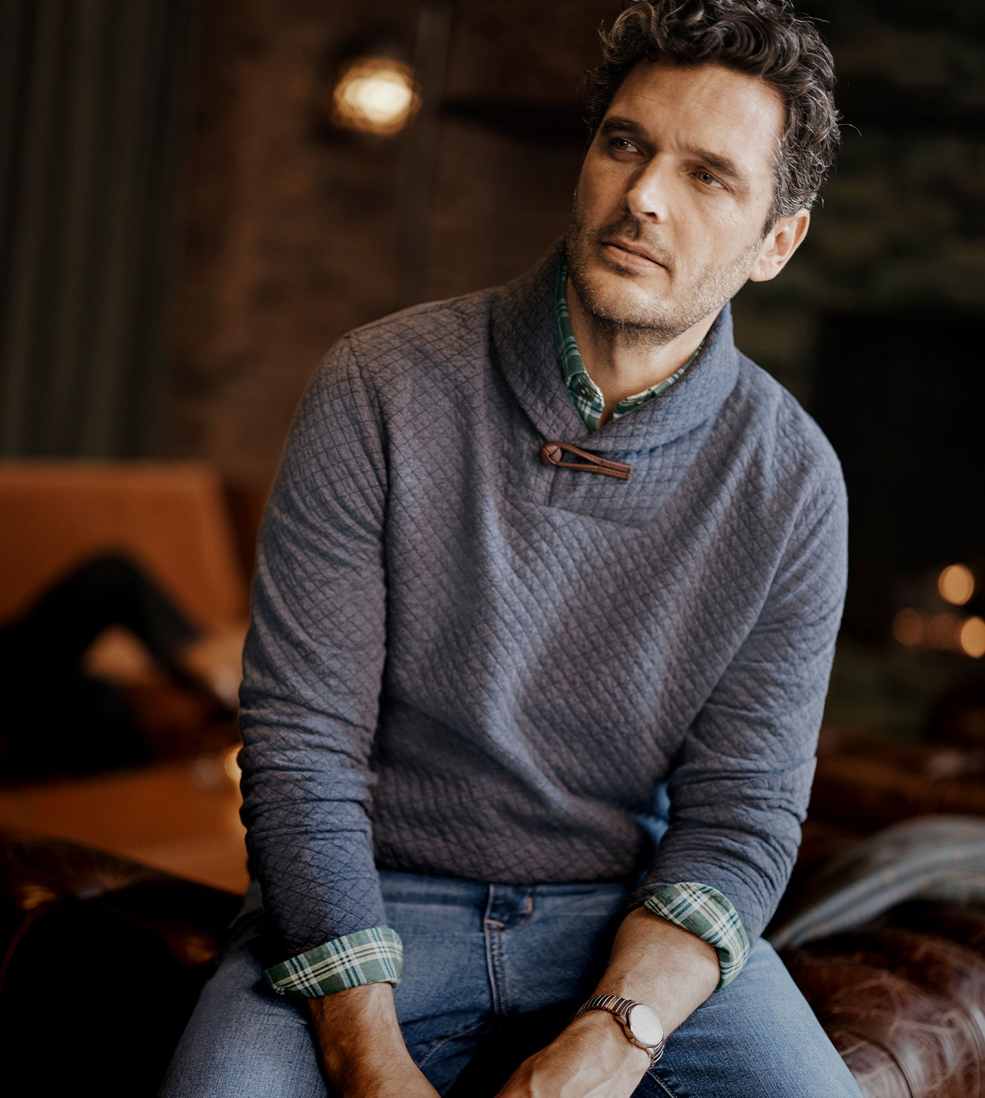 Man sitting on a couch, wearing a sweater, looking contemplative.
