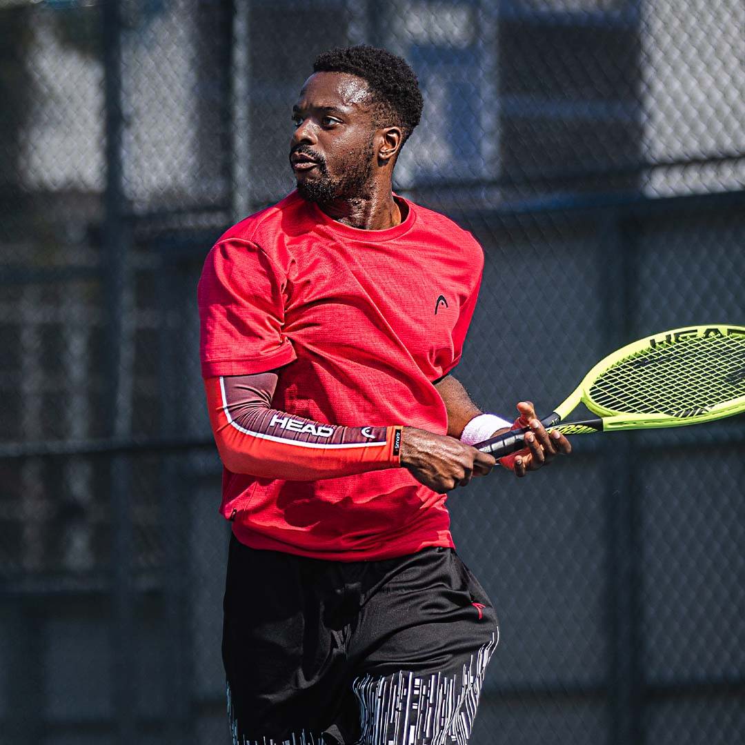 A tennis player pauses mid-game, always keeping his eye on the ball. A smart compression sleeve from HEAD™ is on his arm.